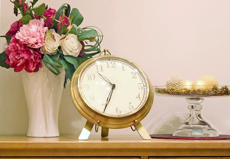 An antique brass tabletop clock sitting on a wooden surface next to a vase of pink and white flowers and a decorative pedaled dish.