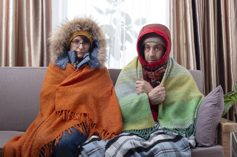 A couple sits shivering on a couch inside their home, wearing heavy hooded winter coats and wrapped in colorful blankets to stay warm.